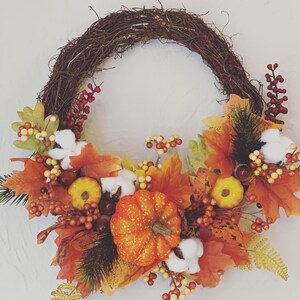May include: A fall-themed wreath with a brown twig base, decorated with orange and yellow pumpkins, red berries, white cotton balls, and orange, yellow, and brown fall leaves.