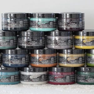Puede incluir: Una disposición piramidal de dieciocho tarros redondos de pintura Terra Clay Paint. Cada tarro cuenta con una tapa negra y una etiqueta con el nombre de la marca, el nombre del color y detalles del producto. Los tarros contienen varios colores de pintura, incluyendo azul, verde, amarillo, rojo y tonos neutros.
