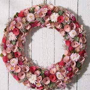 May include: A circular floral wreath with a variety of pink and red flowers, accented with small, light-colored blooms and greenery. The wreath is suspended by a natural twine cord against a white, weathered wooden background.