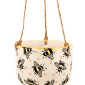 Puede incluir: Un macetero colgante de cerámica blanca con un borde amarillo y un patrón de abejas negras y amarillas. El macetero está suspendido por tres cuerdas de yute marrones.