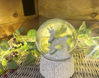 Può includere: Una palla di neve con una figura di renna bianca e dettagli verdi all'interno di una sfera di vetro trasparente. Il globo è posto su una base bianca strutturata. Lo sfondo presenta vegetazione e una superficie intrecciata, creando un'atmosfera festosa.