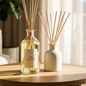 May include: Two reed diffusers on a wooden table. One is a clear glass bottle with a yellow liquid and a label that reads "Citrus Verbena." The other is a small, white ceramic bottle. Both have wooden reeds.
