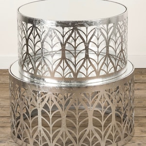 Op de afbeelding: Een set van twee zilveren metalen nesttafels met een geometrisch bladmotief. De bovenste tafel heeft een gespiegelde oppervlak.