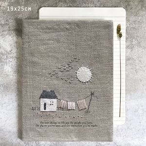 Op de afbeelding: Een grijze linnen notitieboekomslag met een gestikt ontwerp van een huis, een zon en vliegende vogels. De tekst "The best things in life are the people you love, the places you've been and the memories you've made" is gestikt aan de onderkant van de omslag.