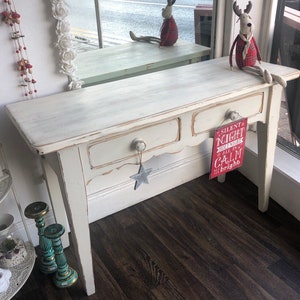 May include: White distressed console table with two drawers and a distressed white finish. The table has a star-shaped ornament hanging from the bottom drawer. A red sign with the text "Silent Night Holy Night All is Calm All is Bright" is hanging from the right side of the table.