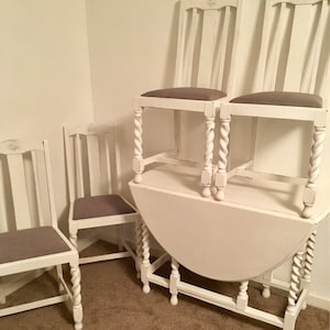 May include: A set of four white chairs with grey cushions and a white round table with a twisted leg design. The table is a drop leaf table and is partially extended.