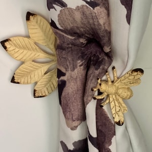 May include: A pair of curtain tiebacks featuring a leaf design and a bee design. The tiebacks are made of metal and have a gold finish.