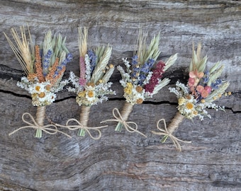 May include: Four dried flower boutonnieres featuring a mix of wheat stalks, purple lavender, white daisies, and colorful foliage. Each small bouquet is tightly wrapped with natural jute twine and finished with a small bow at the base of the stems.