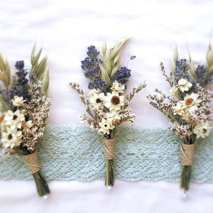 May include: Three boutonnieres featuring dried lavender, white strawflowers, and green oat grass stems. Each bundle is tightly wrapped with natural tan twine at the base, with a small white bead attached to the bottom of the stems.