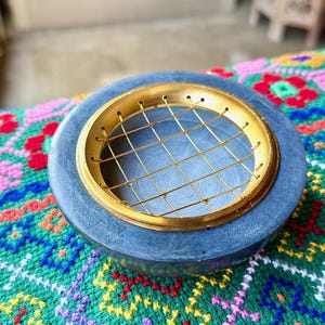 May include: A round, dark gray incense burner with a gold-colored metal grate and rim. The incense burner sits on a colorful, patterned textile. The burner's design is simple and elegant, suitable for home decor.