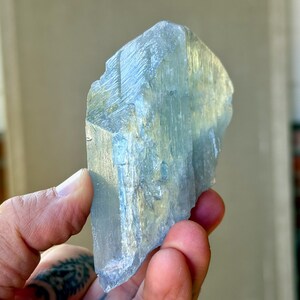 May include: A translucent, light blue crystal with a rough, textured surface. The crystal is held in a hand, showcasing its natural, irregular shape and subtle color variations. The crystal appears to be a raw, uncut gemstone.