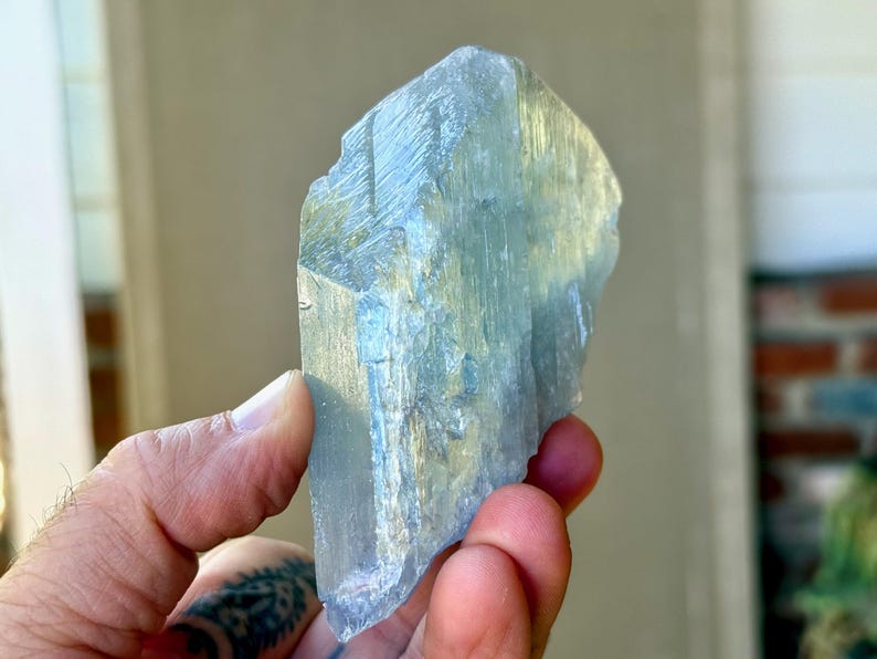 May include: A translucent, light blue crystal with a rough, textured surface. The crystal is held in a hand, showcasing its natural, irregular shape and subtle color variations. The crystal appears to be a raw, uncut gemstone.