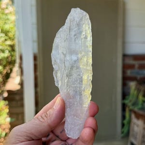 May include: A clear, elongated crystal with a rough, textured surface. The crystal is held in a hand, and the background is blurred, suggesting an outdoor setting. The crystal's shape is irregular, with a natural, raw appearance.