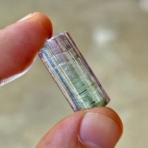 May include: A multi-colored tourmaline crystal held between fingers. The crystal displays a gradient of colors, including pink, green, and clear sections. The gemstone is rectangular with visible striations.