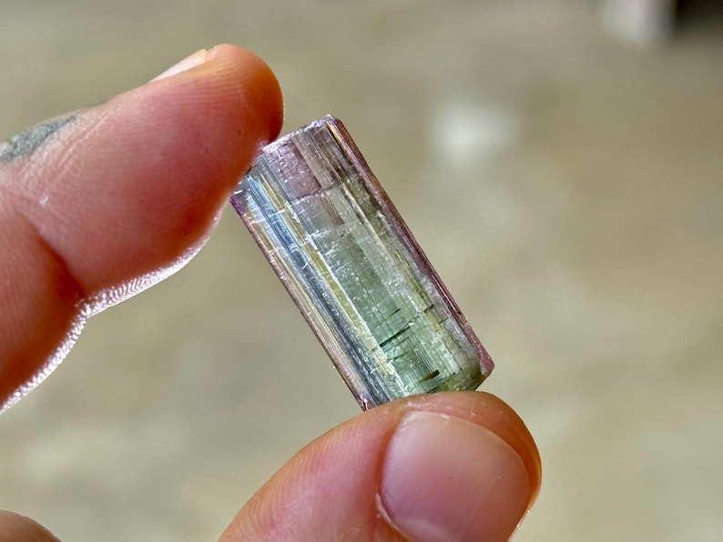 May include: A multi-colored tourmaline crystal held between fingers. The crystal displays a gradient of colors, including pink, green, and clear sections. The gemstone is rectangular with visible striations.