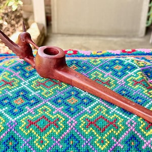 Può includere: Una pipa in legno fatta a mano con un'intaglio di colibrì sulla testa. La pipa è di un ricco colore marrone e ha un lungo e sottile gambo. La pipa è esposta su un tessuto colorato e fantasia.