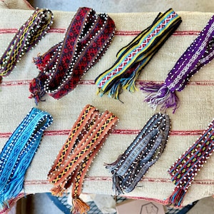Peut inclure: Huit bandes de tissu tissées présentant des motifs géométriques complexes dans diverses couleurs, notamment le rouge, le bleu, le violet, l'orange et le noir. Chaque bande est ornée de petites perles blanches sur les bords et de franges.