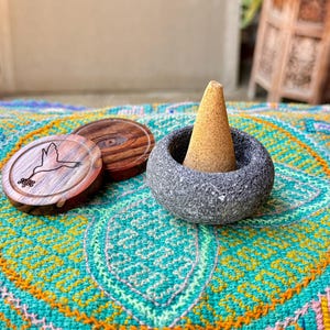 May include: A grey stone incense burner with a beige incense cone inside. The burner is sitting on a colourful patterned rug with two wooden coasters with carved designs.