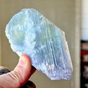 May include: A large, raw, blue-green crystal with a layered, textured appearance. The crystal is held up, showcasing its natural form and color variations. The stone has a translucent quality, with visible striations and a rough, uneven edge.