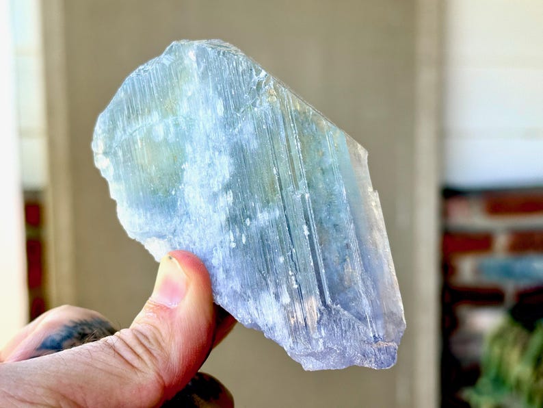 May include: A large, raw, blue-green crystal with a layered, textured appearance. The crystal is held up, showcasing its natural form and color variations. The stone has a translucent quality, with visible striations and a rough, uneven edge.