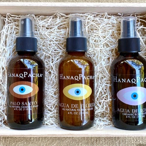 May include: Three brown glass bottles with black spray tops in a wooden box filled with shredded paper. Each bottle has a white label with the text "HANAQPACHA" and a blue eye with a different colored background. The bottles are labeled "Palo Santo", "Agua de Florida", and "Agua de Rosas".