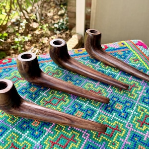May include: Four handcrafted wooden pipes with a dark brown finish are arranged on a colorful, patterned textile. The pipes have a curved design with a bowl and a long, tapered stem. The pipes are likely for smoking.