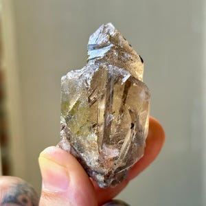 May include: A raw, natural crystal cluster with a translucent, smoky quartz appearance. The crystal has a rough, textured surface with varying shades of brown and gray. The crystal is held in a hand, showcasing its natural form and unique characteristics.