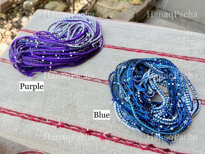 K&ouml;nnte beinhalten: Zwei Garnrollen, eine lila und eine blaue, mit wei&szlig;en Perlen, die in das Garn eingewebt sind. Die Rollen liegen auf einer gestreiften Stoffoberfl&auml;che. Der Text "Purple" befindet sich unter der lila Rolle und der Text "Blue" unter der blauen Rolle.
