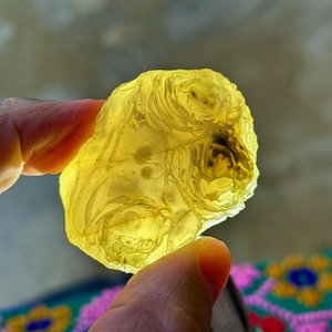 May include: A translucent, irregularly shaped, yellow-toned stone is held between fingers. The stone has a textured surface with internal patterns and a darker, more opaque area. The light shines through the stone, highlighting its details.