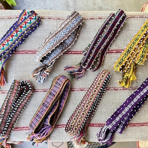 May include: An assortment of eight colorful woven belts with intricate patterns and beaded accents. Each belt features a unique design with tassels at the ends. The belts are displayed on a neutral-toned, striped surface.
