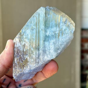 May include: A large, raw, blue-green kyanite crystal held in a hand. The crystal has a layered, fibrous appearance with streaks of gold and a rough, uneven texture. The stone is a natural mineral specimen.