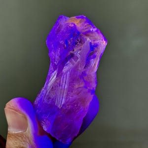 Może przedstawiać: Zbliżenie surowego, krystalicznego okazu mineralnego. Minerał ma żywy fioletowy kolor z obszarami pomarańczowego i żółtego. Widoczna jest struktura krystaliczna, z ostrymi krawędziami i szorstką fakturą. Tło jest stonowane szaro-zielone.