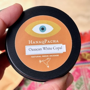May include: A black, round tin of HanaqPacha Oaxacan White Copal natural resin incense. The label features an eye design and a hummingbird illustration. The tin is held in a hand.