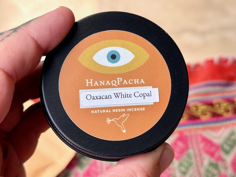 May include: A black, round tin of HanaqPacha Oaxacan White Copal natural resin incense. The label features an eye design and a hummingbird illustration. The tin is held in a hand.
