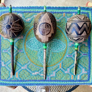 May include: Three handcrafted gourd rattles with intricate etched designs. The gourds are dark brown with white and tan patterns, attached to wooden handles with green accents. They rest on a blue and green patterned textile.