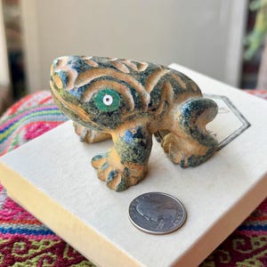 Puede incluir: Una figurita de rana tallada de color verde y amarillo con un ojo blanco y verde. La rana está sentada en una caja blanca con una moneda a su lado.