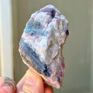 May include: A raw mineral specimen featuring a combination of white quartz and dark blue-green tourmaline crystals. The stone has a rough, uneven texture with visible vertical striations and patches of deep magenta-colored lepidolite inclusions.