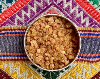 May include: A close-up of a metal container filled with small, translucent, amber-coloured resin pieces. The container is sitting on a colourful woven fabric.