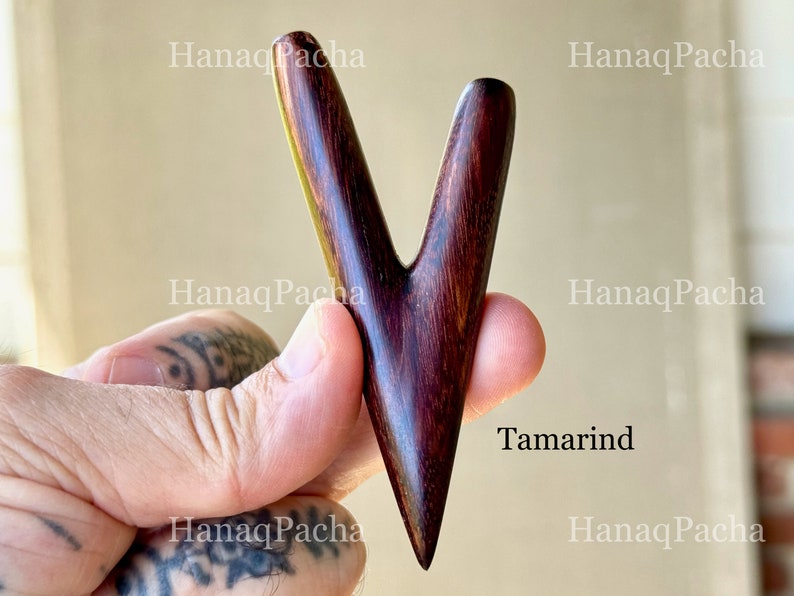 May include: A V-shaped massage tool crafted from dark brown tamarind wood with a smooth, polished finish. The object features two tapered prongs extending from a single pointed base, showcasing the natural grain patterns of the wood throughout.