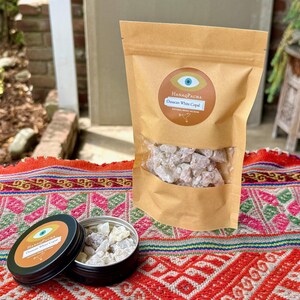 May include: A brown paper bag and a small black tin, both labelled "HanaqPacha Oaxaca White Copal", are on a colourful textile. The bag contains white copal resin, and the tin is open, revealing more of the resin.