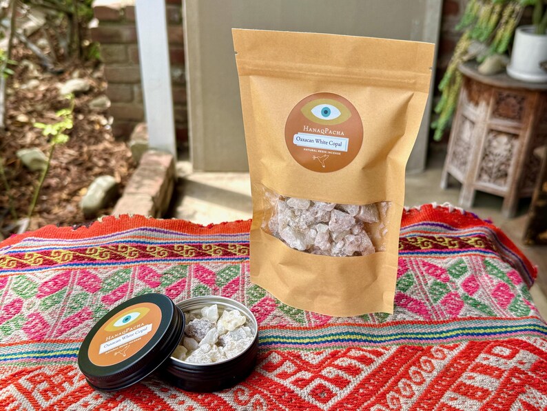 May include: A brown paper bag and a small black tin, both labelled "HanaqPacha Oaxaca White Copal", are on a colourful textile. The bag contains white copal resin, and the tin is open, revealing more of the resin.