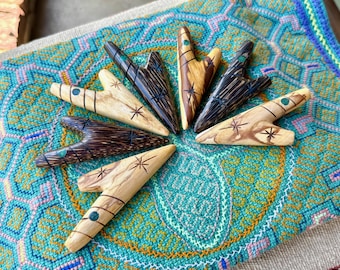 May include: Six wooden arrow-shaped ornaments with green gemstone accents. The ornaments are arranged in a circle on a colorful patterned fabric.