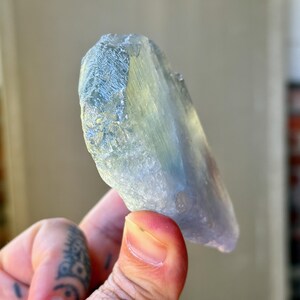 May include: A translucent, light blue crystal with a textured surface. The crystal has a natural, irregular shape and is held in a hand. The crystal's color shifts from blue to a pale yellow-green.