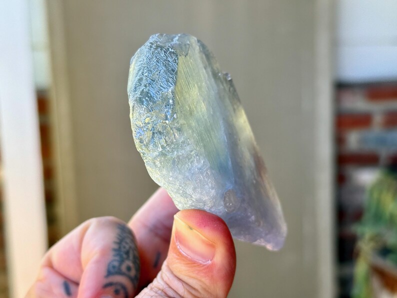 May include: A translucent, light blue crystal with a textured surface. The crystal has a natural, irregular shape and is held in a hand. The crystal's color shifts from blue to a pale yellow-green.