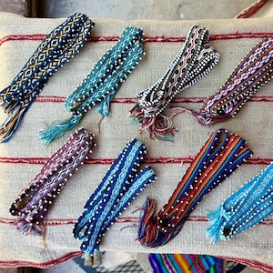 May include: Eight woven fabric bands featuring intricate geometric patterns in various colors including blue, teal, black, red, and white. Each band is embellished with small white beads along the edges and finished with frayed tassel ends.