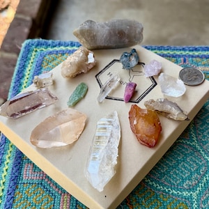 May include: A collection of various raw mineral crystals and gemstones arranged on a square beige cardboard box. The box features a black hexagonal logo with a hummingbird graphic. A silver coin is placed on the right side of the box surface.