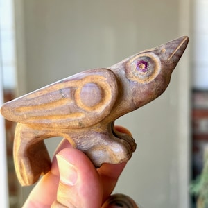 May include: A small, hand-carved wooden bird figurine with a smooth, light brown finish. The bird features stylised wing carvings, a pointed beak, and a circular eye detail with a small, dark purple inlay at the centre of the eye socket.