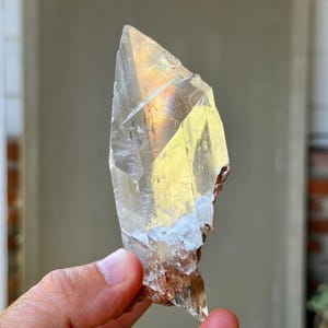 以下が含まれることがあります： 尖った上部とざらざらした質感の基部を持つ透明な水晶。クリスタルは半透明で、黄色と茶色の色合いがあります。光がファセットを捉え、きらめく効果を生み出しています。クリスタルは手で持たれています。