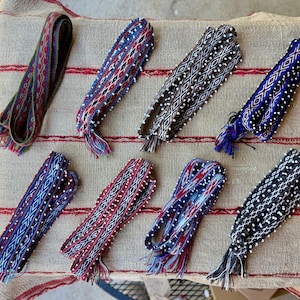 May include: Eight woven fabric bands featuring intricate geometric patterns in various color combinations including red, blue, black, and white. Several of these bands are embellished with small white beads along the edges and have fringed ends.