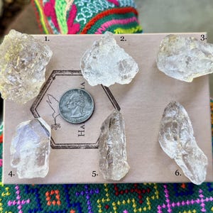 May include: Six raw, clear quartz crystal specimens of varying shapes and sizes are displayed on a cardboard surface. A US quarter coin is included for size reference. The crystals are numbered 1 through 6. The background includes colorful woven textiles.